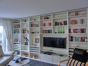 Uma sala de estar com uma televisão de tela plana grande, estantes brancas com livros e objetos decorativos, um sofá e uma mesa de centro.
