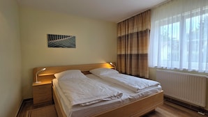 Een netjes opgemaakt bed met witte lakens, een nachtkastje met een lamp, een radiator en een raam met dunne gordijnen.
