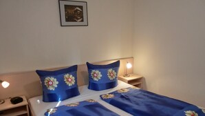 Una cama bien hecha con ropa de cama azul y almohadas estampadas, una lámpara de noche y un pequeño estante con una radio.