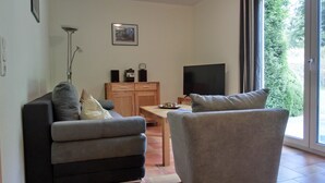Una sala de estar con un sofá gris, un sillón gris, un mueble de madera con un televisor de pantalla plana y un ventanal con vistas al follaje.