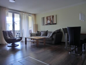 Ein Wohnzimmer mit einer Couch, einem Couchtisch, einem Sessel und einem Esstisch. An der Wand hängt ein Gemälde und ein Fenster mit Vorhängen.