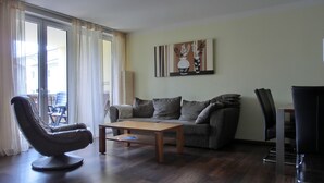 A modern living room with a sofa, a coffee table, a chair, and a dining table. There is a large window with curtains, a painting on the wall, and a view of the outside. - 3-zi.-fewo für 4+2 Personen - mit Telefon- und Internetanschluss (Graal-Müritz)