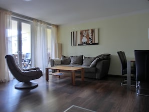 Un salon moderne comprenant un canapé, une table basse, un fauteuil et une table à manger. Il y a une grande fenêtre avec des rideaux, un tableau au mur et une vue sur l’extérieur.