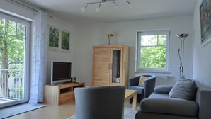 Moderni olohuone, jossa on harmaa sohva, puinen kaappi, televisio puisen jalustan päällä ja suuri ikkuna, josta avautuu näkymä vehreään maisemaan.