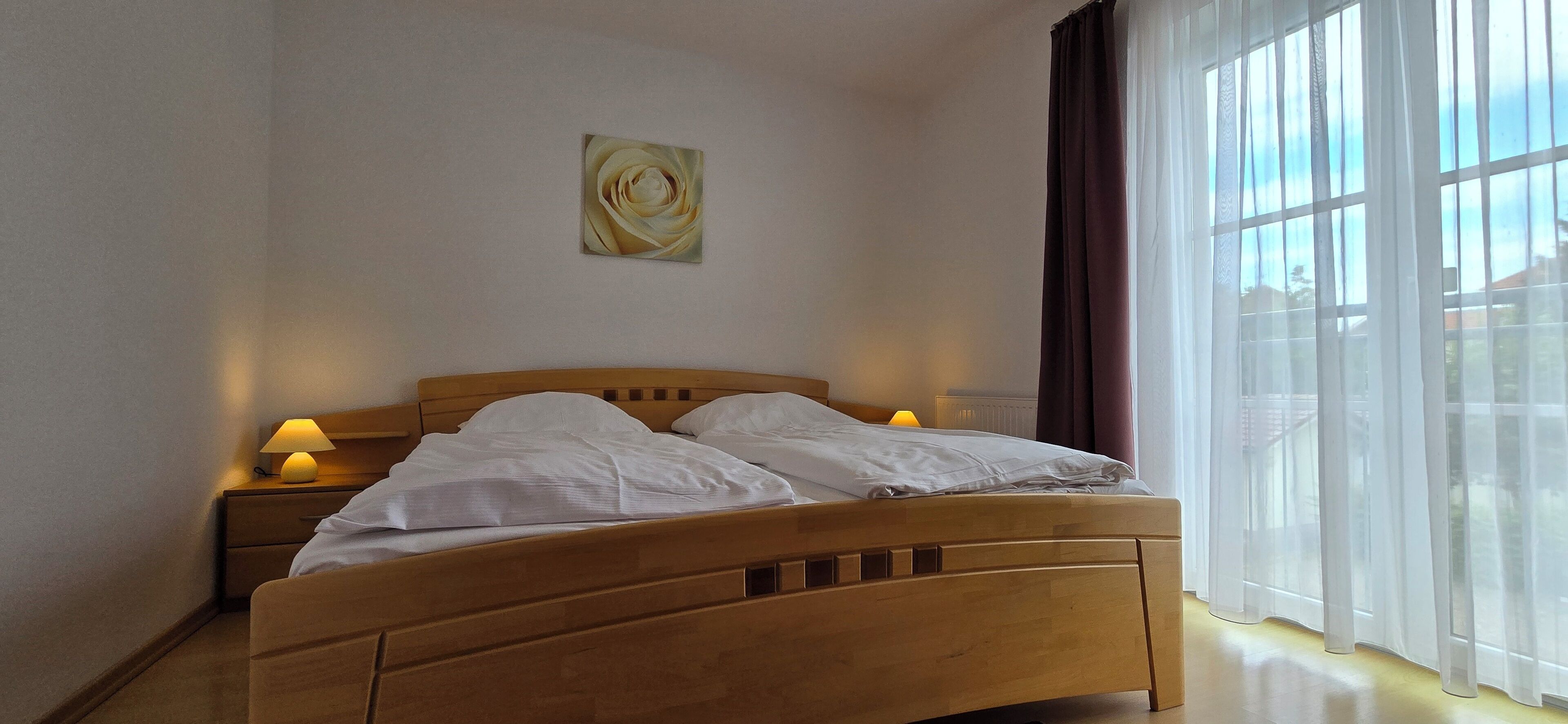 Quarto com cama de madeira, duas luminárias de cabeceira, janela com cortinas finas e um quadro na parede.