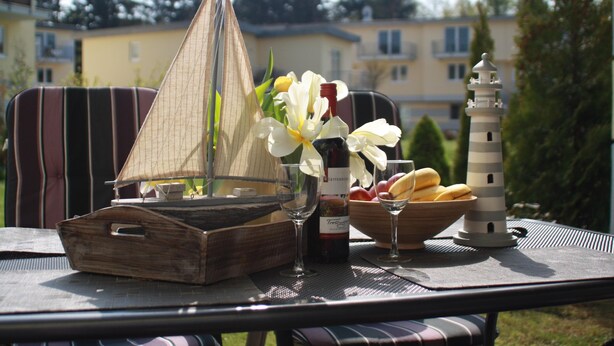 Une table sur laquelle on trouve une maquette de voilier, une bouteille de vin, un verre à vin, un panier de fruits et une maquette de phare.
