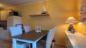 Un coin repas avec une table en bois et des chaises blanches, une petite décoration représentant un voilier accrochée au mur, et une lampe posée sur une console blanche.