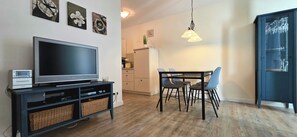 A living room with a flat-screen TV, a dining table with chairs, a kitchen area, and a cabinet with glassware. - Strandnahe 2-zimmer-fewo im 1. OG für 3 Pers (Graal-Müritz)
