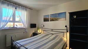 Ein Schlafzimmer mit Bett, einem Fenster mit Vorhängen, einer Heizung, einem Nachttisch mit Lampe und einem an der Wand montierten Fernseher.
