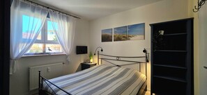 Une chambre à coucher avec un lit, une fenêtre avec des rideaux, un radiateur, une table de chevet avec une lampe et un téléviseur fixé au mur.
