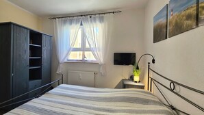 Et soveværelse med seng, garderobeskab, et vindue med gardiner, en radiator, et natbord med lampe og et TV ophængt på væggen.