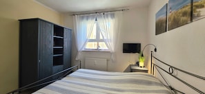 A bedroom with a bed, a wardrobe, a window with curtains, a radiator, a bedside table with a lamp, and a wall-mounted TV. - Strandnahe 2-zimmer-fewo im 1. OG für 3 Pers (Graal-Müritz)