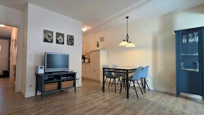 Ein modernes Wohnzimmer mit einem Flachbildfernseher, einem Essbereich mit blauen Stühlen und einer Küche im Hintergrund.