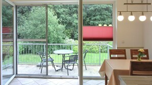 A room with a sliding glass door leading to a balcony with a table and chairs, a dining area with a table and chairs, and a hanging light fixture.