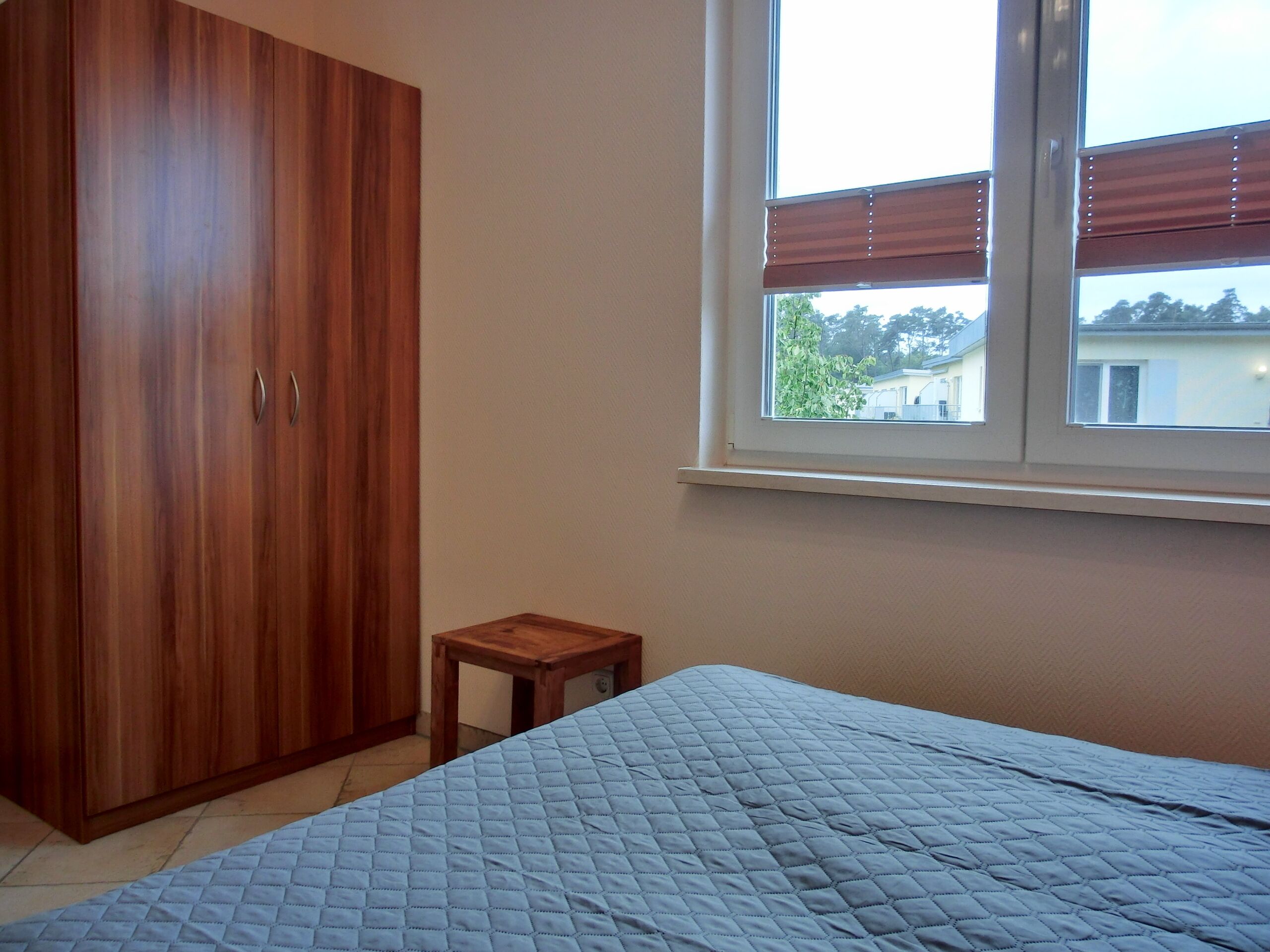 A bedroom with a bed, a wooden wardrobe, a small wooden table, and a window with blinds.