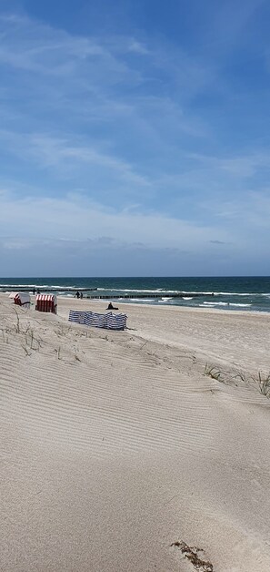 Une plage de sable avec un parasol rayé, un abri de plage rayé de rouge et de blanc, et une jetée en bois qui s’avance dans l’eau.