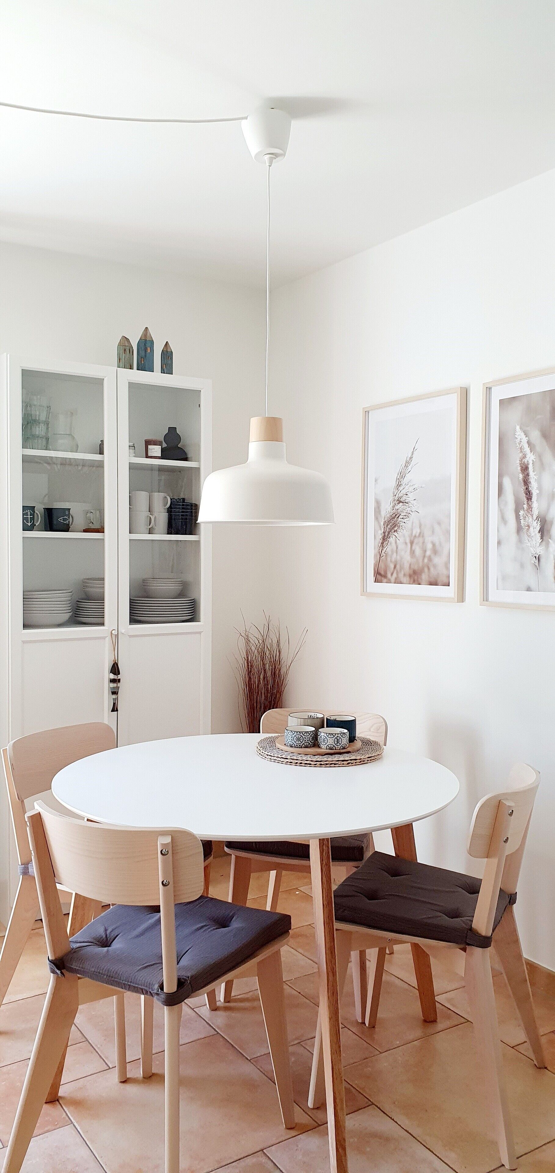 A dining area with a round white table, four wooden chairs, a white pendant light, and a cabinet with glass doors in the background.