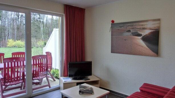 Een kamer met een schuifdeur naar een tuin, een flatscreen televisie op een klein tafeltje, een rode bank en een schilderij van een strand.