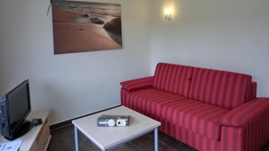 Een woonkamer met een rood gestreepte bank, een flatscreen televisie op een standaard, een kleine tafel met een afstandsbediening en een boek, en een wanddecoratie met een strandthema.