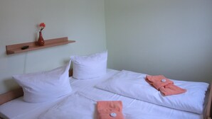 Een kamer met twee bedden, witte beddengoed, een houten plankje met een vaas en een afstandsbediening, en een effen muur.