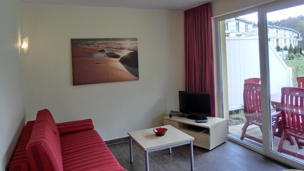 A room with a red sofa, a small table, a TV on a stand, and a large window with a view of buildings and greenery. - 2-zimmer-fewo für 5 Pers. EG mit Terrasse (Graal-Müritz)