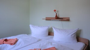 A bed with white bedding and pillows, a pink towel, and a small vase with a flower on a wall-mounted shelf. - 2-zimmer-fewo für 5 Pers. EG mit Terrasse (Graal-Müritz)