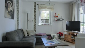 Een woonkamer met een grijze bank, een salontafel met tijdschriften, een televisie op een houten standaard en een raam met gordijnen.