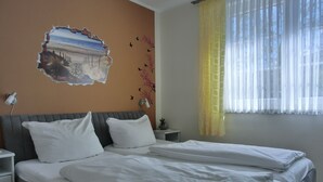 Een tweepersoonsbed met wit beddengoed, een grijs hoofdbord, een nachtkastje met een lamp en een wanddecoratie van een strandgezicht.