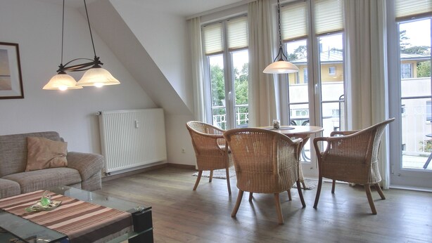 Un salon moderne comprenant un canapé, une table basse en verre, des fauteuils en osier et une table à manger. De grandes fenêtres laissent entrer la lumière naturelle et offrent une vue sur l’extérieur.