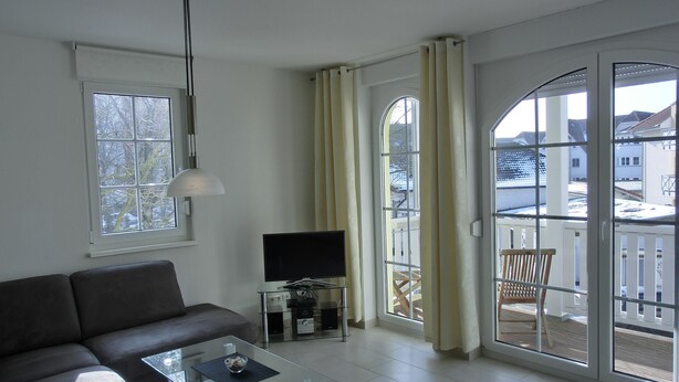 A modern living room with a sofa, a glass coffee table, a TV, and a balcony with curtains. - 3-zimmer-fewo für 4 Pers. im 1. OG (Graal-Müritz)