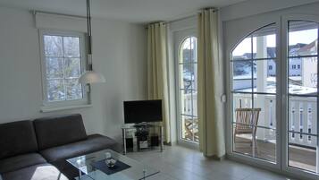 Ein modernes Wohnzimmer mit einer Couch, einem Glastisch, einem Fernseher und einem Balkon mit Vorhängen.