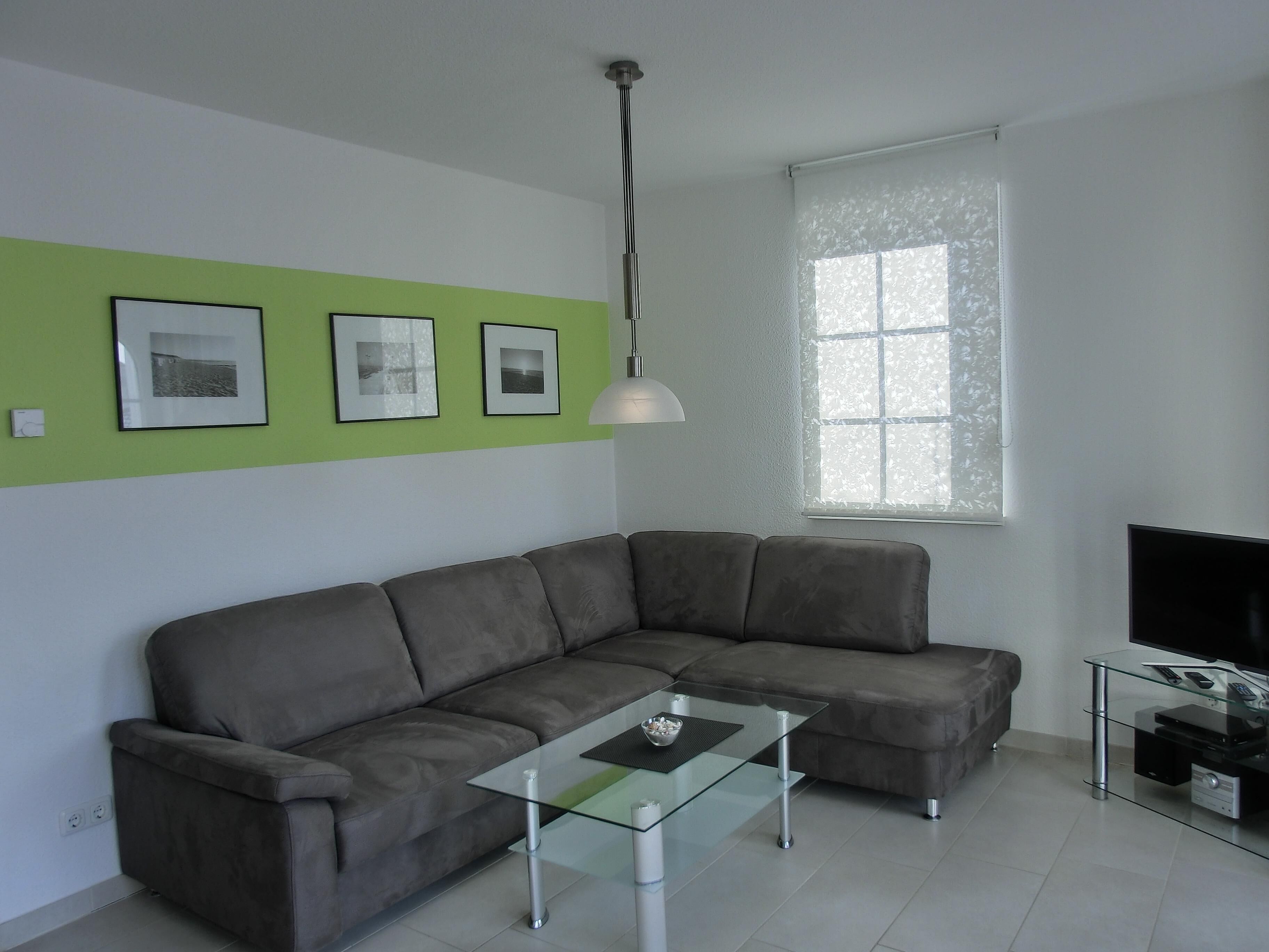 Ein modernes Wohnzimmer mit einem grauen Ecksofa, einem Glastisch, einem Flachbildfernseher auf einem TV-Lowboard aus Glas und einem Fenster mit Milchglas.