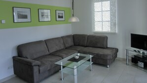 A modern living room with a grey sectional sofa, a glass coffee table, a flat-screen TV on a glass TV stand, and a window with frosted glass. - 3-zimmer-fewo für 4 Pers. im 1. OG (Graal-Müritz)