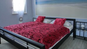 Um quarto com uma cama, uma janela com cortinas, uma obra de arte na parede retratando uma praia e uma mesa de cabeceira com luminária.