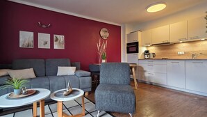 A modern living room with a grey sofa, a grey armchair, and a small table with a plant. A kitchen with white cabinets and a wooden countertop is visible in the background. - 2-zi.-fewo im 2. OG, ca. 45 m² für 4 Personen mit Wlan (Graal-Müritz)