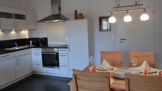 A modern kitchen with white cabinets, a built-in oven, and a black countertop. There is a dining table with chairs and a chandelier above. - 2-zi.-fewo für 4 Personen im 2. OG (Graal-Müritz)