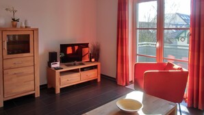 A living room with a wooden cabinet, a TV on a wooden stand, a red chair, and a large window with curtains. - 2-zi.-fewo für 4 Personen im 2. OG (Graal-Müritz)