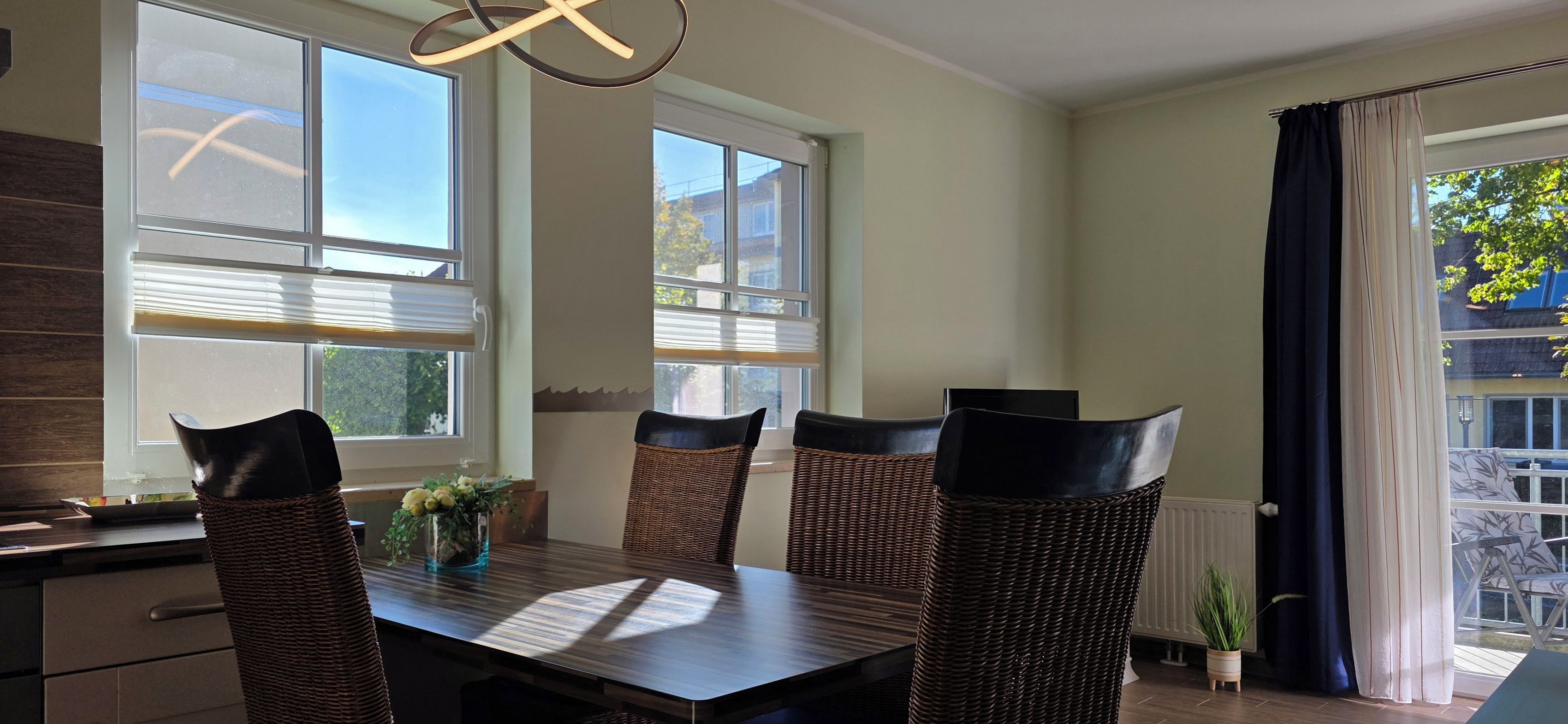 Un comedor con una mesa de madera oscura y cuatro sillas de mimbre con cojines negros. Hay dos ventanas con persianas, y una vista al exterior por la ventana de la derecha.