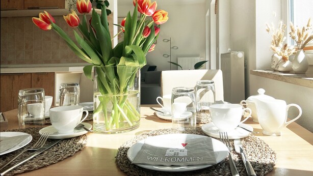 Ein Esstisch ist gedeckt mit weißen Tellern, Besteck und Gläsern, in der Mitte steht ein Strauß Tulpen.