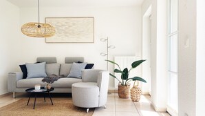 Ein modernes Wohnzimmer mit grauem Sofa, rundem Ottomanen, Couchtisch und einer hängenden Lampenschirm aus Weidengeflecht. Ein großes Fenster sorgt für natürliches Licht, und eine Topfpflanze steht in der Ecke.