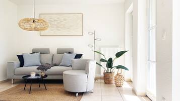 Ein modernes Wohnzimmer mit grauem Sofa, rundem Ottomanen, Couchtisch und einer hängenden Lampenschirm aus Weidengeflecht. Ein großes Fenster sorgt für natürliches Licht, und eine Topfpflanze steht in der Ecke.