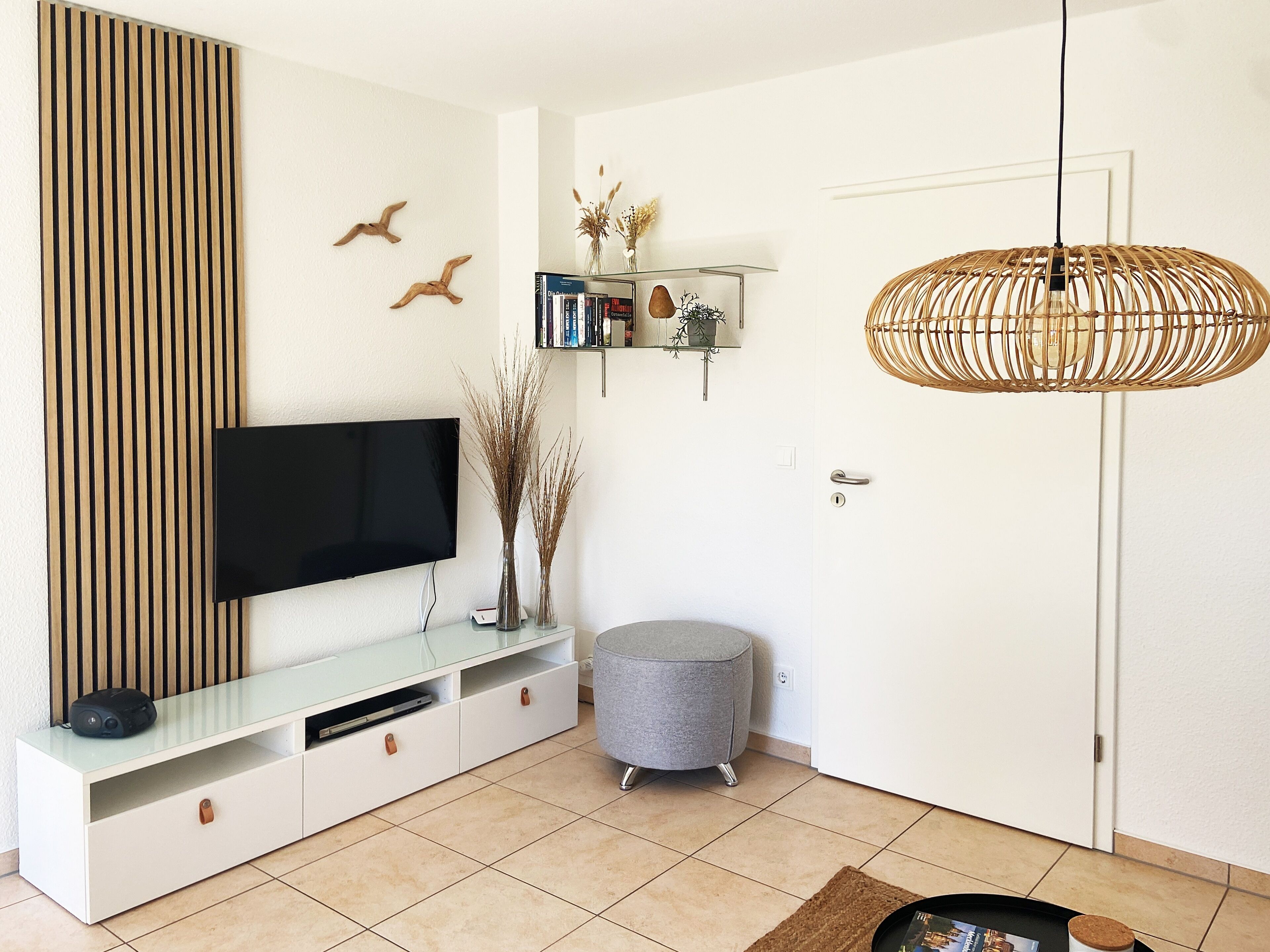 Un salon moderne doté d’un téléviseur à écran plat, d’un meuble TV en verre, d’un pouf gris, d’une suspension en rotin et d’une porte à serrure à clé.