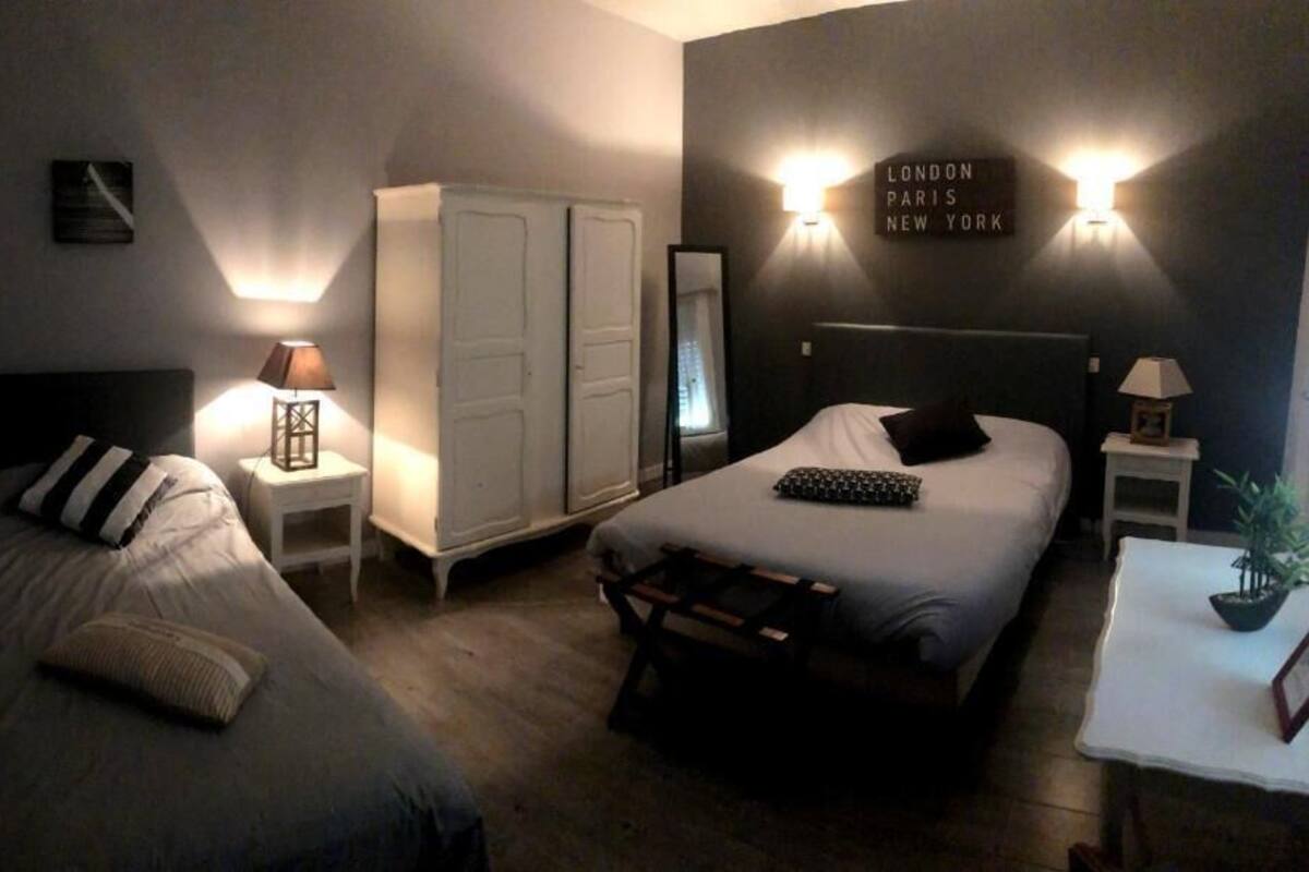 A hotel room with two beds, a wardrobe, bedside tables with lamps, and a wall sign listing London, Paris, and New York.