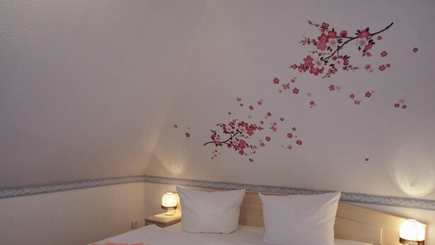Una cama bien tendida con sábanas blancas y toallas rosas, una pared con un mural floral y lámparas de noche sobre una mesita.