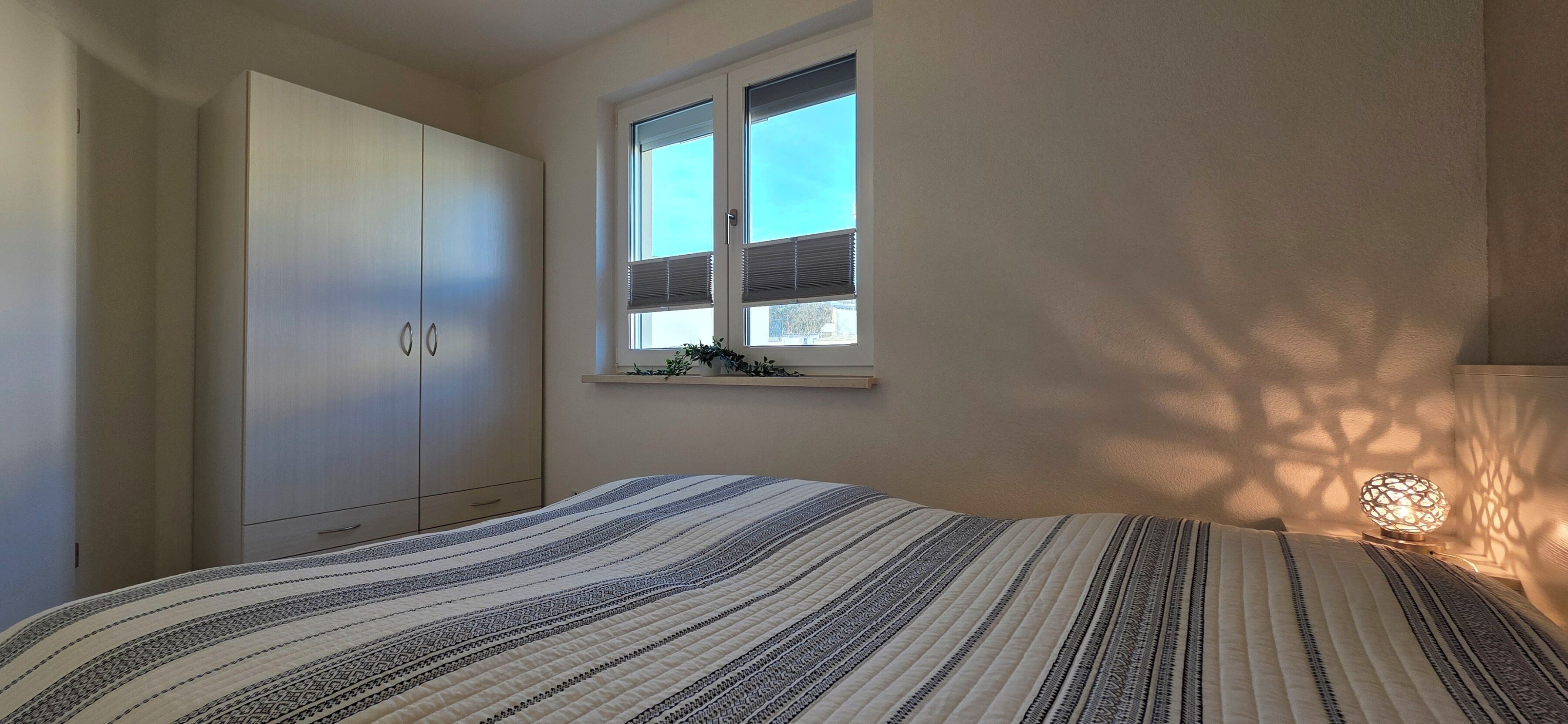 Quarto com cama, guarda-roupa, janela com persianas e luminária fixada na parede.