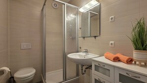 Ein modernes Badezimmer mit einer Duschkabine aus Glas, einem weißen Waschbecken und einem Spiegel darüber. Auf der Ablage steht ein Blumentopf und auf der Arbeitsfläche ein aufgerolltes Handtuch.