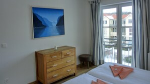 A hotel room with a bed, a wooden dresser, a small stool, a painting of a fjord, and a window with curtains.