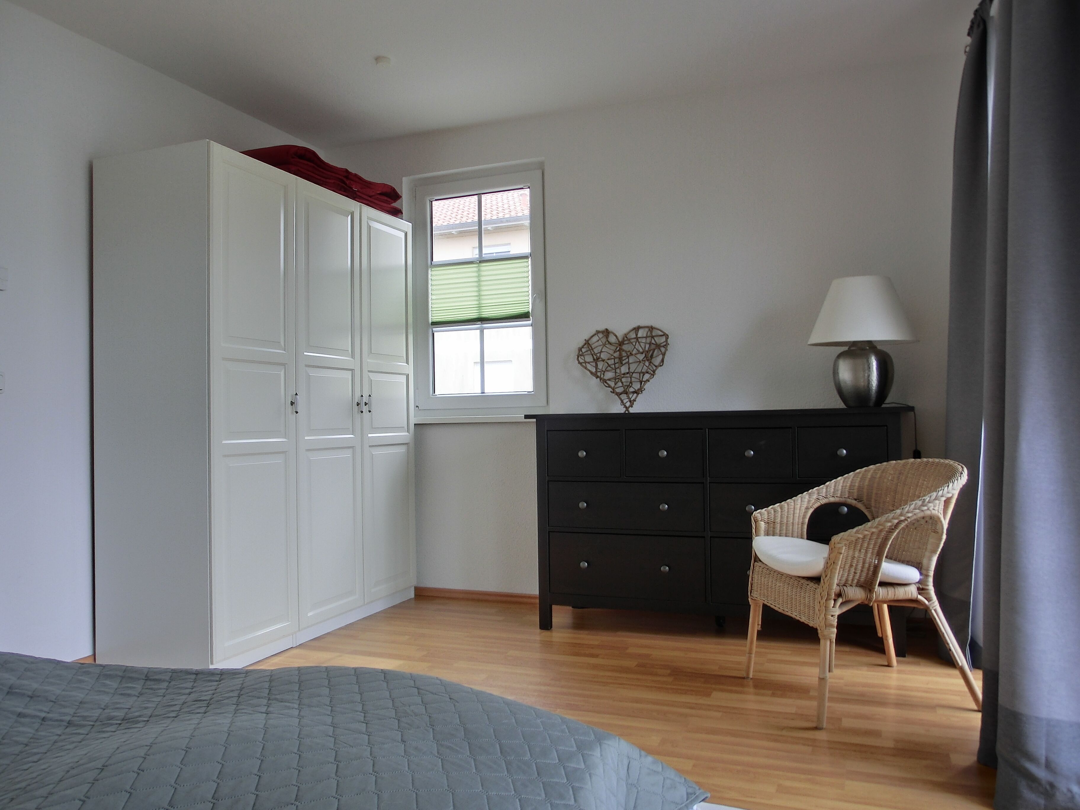 A bedroom with a bed, a dresser, a chair, a window, and a heart-shaped decoration on the wall.