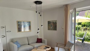 A modern living room with a sofa, coffee table, and chair, a painting on the wall, and a view through a glass door. - 3-zi.-fewo in Angenehmer und Ruhiger Lage im Erdgeschoss für 6 Pers (Graal-Müritz)