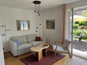 Ein modernes Wohnzimmer mit Sofa, Couchtisch und Sessel, einem Bild an der Wand und Blick durch eine Glastür.